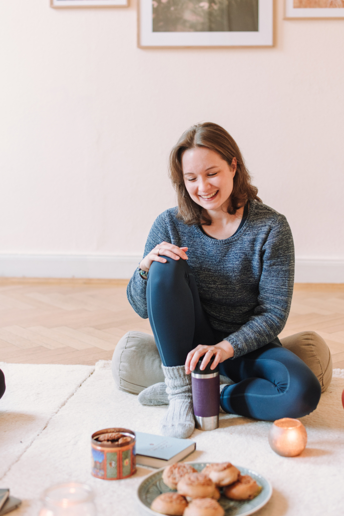 Nina sitzt in einem blauen Pullover auf einem Yogakissen und isst Zimtschnecken